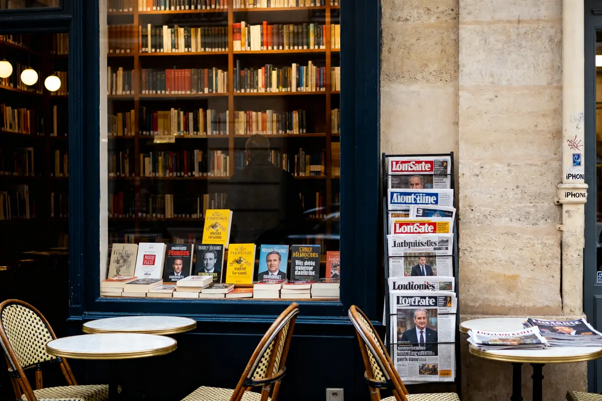 Devanture de librairie avec journaux et livres exposés près d'une table café score IAE Message.